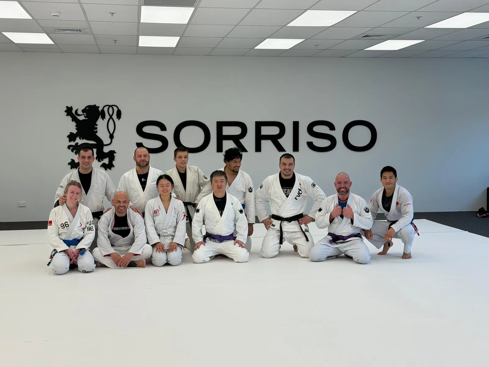 An adults class at Sorriso BJJ Botany gathered on the white mats in front of the Sorriso logo on the far wall